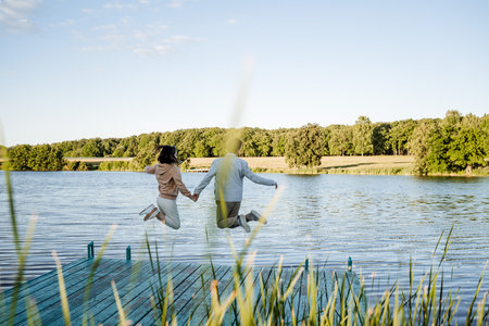 A man and a woman are joyfully jumping into a beautiful lake while holding hands tightly, surrounded by natures beauty and greeneryの写真素材