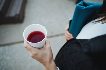 A stylish woman confidently holding a cup filled with a dark red drink, while carrying important documents tucked under her arm, enjoying the outdoors around herの写真素材