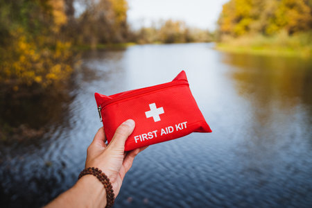 A person is holding a first aid kit in front of a serene body of water, surrounded by natural beauty and vibrant scenery. This scene captures a moment of preparedness amidst a peaceful landscapeの写真素材