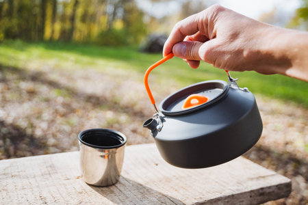 Enjoying the outdoors, a hand pours hot water from a kettle into a stainless steel cup on a wooden surface surrounded by natureの写真素材