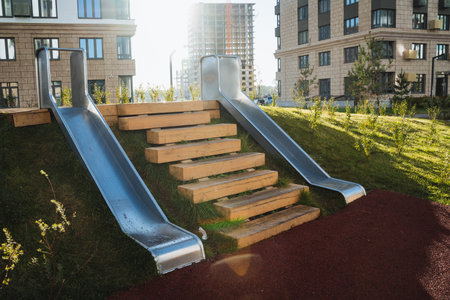 In a landscaped park, a modern metal slide and sturdy wooden steps ascend a gentle hill, inviting families to enjoy outdoor activities, promoting engagement with nature and relaxationの写真素材