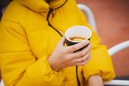 A woman, who is wearing a bright yellow jacket, is holding a steaming cup of coffee in her hand, enjoying a moment of warmth and comfort. The drink is a delightful beverage that she savorsの写真素材