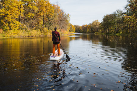 A paddleboarder glides on a calm river, surrounded by vibrant autumn foliage and shimmering reflections, perfect for themes of adventure and outdoor explorationの写真素材