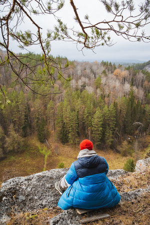 A person is comfortably seated on a large, sturdy rock that provides a stunning, panoramic view of a lush green forest, completely surrounded by the serene beauty and tranquility of natureの写真素材