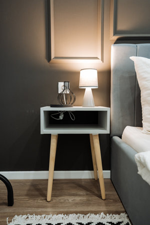 In a cozy bedroom, there is a stylish nightstand adorned with a charming lamp, positioned conveniently next to a comfortable bed, creating a warm and inviting atmosphere for relaxationの写真素材