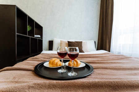 On a comfortable bed, there are two elegantly positioned glasses of wine along with two freshly baked croissants, beautifully arranged on a decorative tray. This setting creates a cozy atmosphereの写真素材