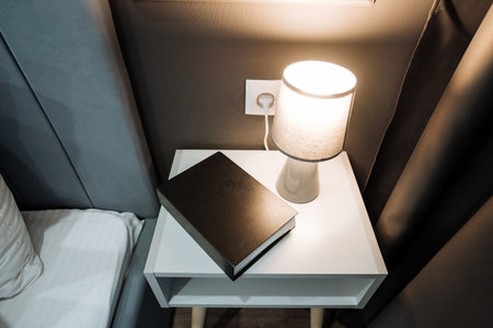 In the softly lit room, a beautifully arranged book lies gracefully on the nightstand, accompanied by a lamp that emits a warm, inviting glow, enhancing the overall coziness and charm of the spaceの写真素材