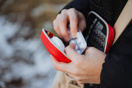 A person is carefully taking a small pill out of a vibrant red case, which holds it securely,...