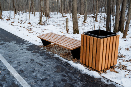 A sturdy wooden bench, complemented by a trash can, sits quietly on the side of a snowcovered road,...
