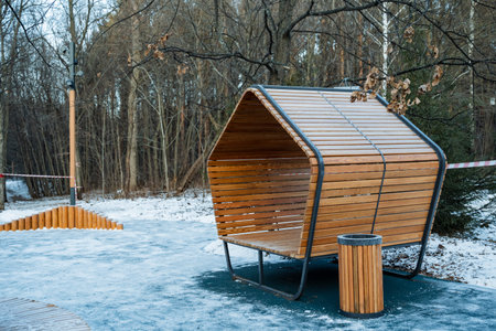 In a winter wonderland, a charming wooden bench sits beside a trash can in a snowy park, creating a picturesque scene amidst the serene beauty of nature and the falling snowflakesの写真素材