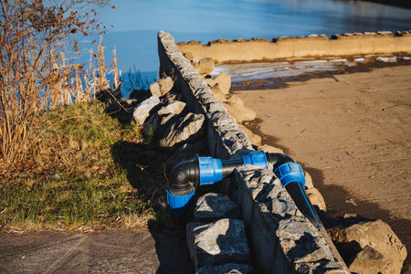 A vibrant blue pipe has been securely attached to a sturdy stone wall that is positioned right next to a calm body of water, creating an interesting visual scene for onlookersの写真素材