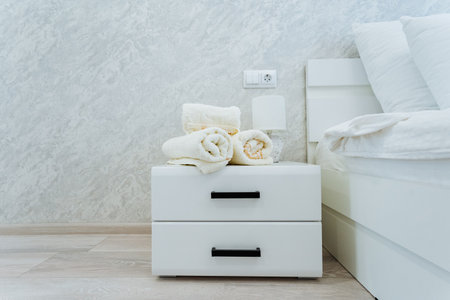 A contemporary white nightstand positioned next to a snug bed, featuring neatly arranged plush towels on top, elevates the rooms overall ambiance, fostering a warm and inviting atmosphereの写真素材