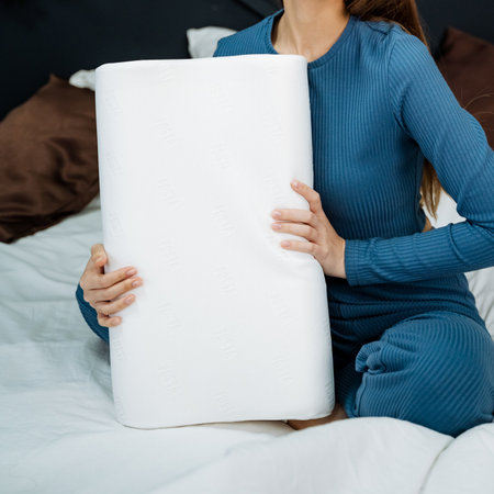 A woman sits comfortably on a cozy bed, cradling a fluffy white pillow. This serene setting enhances her tranquility, allowing her to relax and unwindの写真素材