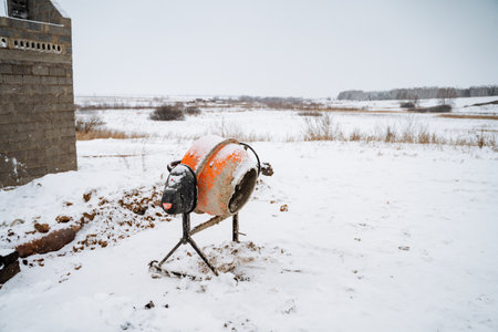 A heavyduty concrete mixer sits motionless in thick snow outside a sturdy building, surrounded by a stunning winter landscape, highlighting icy conditions and frigid temperaturesの写真素材
