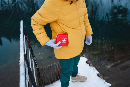 A person dressed in a bright yellow jacket is standing by a snowy lake while holding a first aid kit. This scene radiates adventure and caring amidst a beautiful winter landscapeの写真素材