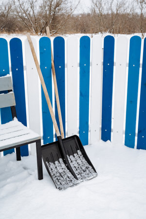 Two shovels are resting in the snow, right in front of a lovely blue and white fence that stands against the frosty winter backdrop, creating a picturesque and serene sceneの写真素材
