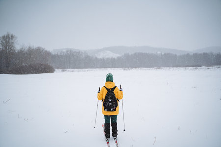 A person clad in a yellow jacket skis across a snowy field, with snow-covered mountains visible in the distance under an overcast skyの写真素材