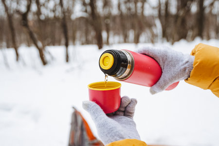 Person in gloves pours warm beverage from thermos into red cup amidst snowy landscape, surrounded by trees, emphasizing cozy winter vibesの写真素材