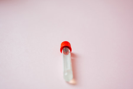 A laboratory test tube featuring a bright red cap is currently positioned on a soft pink surface,...