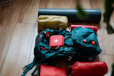 On a wooden floor, there is a distinctive backpack filled with a bright red first aid kit, which is...