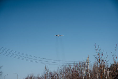 An airplane is flying through a clear blue sky above a landscape with trees and power lines.