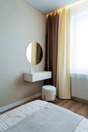 This furnished bedroom offers a cozy bed, an elegant ottoman for seating, a stylish mirror for decor, and a uniquely designed window that lets in natural light, creating a warm atmosphereの写真素材