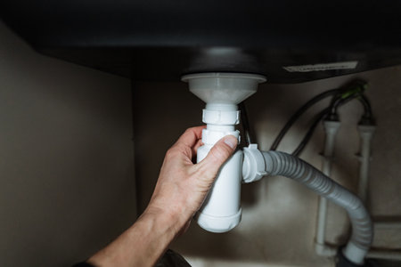 An individual is diligently fixing a sink pipe that is positioned beneath a kitchen sink to ensure...