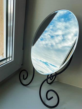 A beautifully crafted round mirror is positioned on a window sill, reflecting its stunning surroundings with an enchanting charm that captivates anyone who gazes at its exquisite surfaceの写真素材