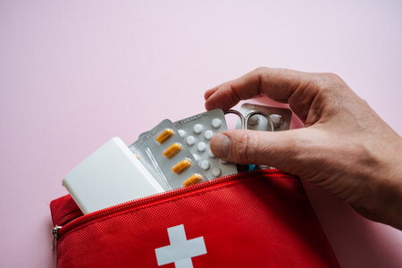 A closeup of a hand retrieving medication from a red first aid kit, highlighting health and wellness concepts against a soft pink backdrop that conveys caring and safetyの写真素材