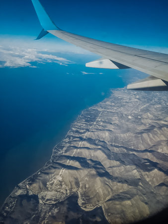 The wing of an airplane is soaring high while flying gracefully over the vast ocean and the majestic mountains below. This amazing journey showcases the beauty of air travel and natureの写真素材