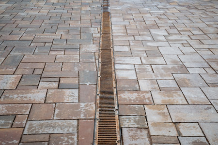 There is a drain situated between two bricks along the path of a sidewalk, where pedestrians walk, providing a functional means for water drainage and maintaining the integrity of the walkwayの写真素材