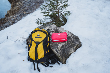 In the midst of a breathtaking snowy landscape, there rests a vivid bright yellow backpack alongside a striking red first aid kit, both thoughtfully positioned on a large, snowcovered rock nearbyの写真素材