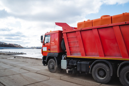 A bright red dump truck stops by a serene body of water, creating a picturesque scene that blends natures beauty with human engineering, captivating all who take a moment to observeの写真素材
