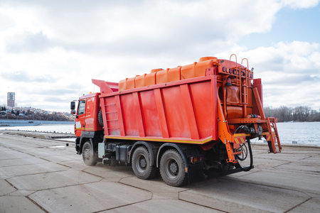 A bright red dump truck is parked conveniently and neatly in a spacious parking lot, which is situated next to a large, beautiful body of water that perfectly reflects the sunlightの写真素材