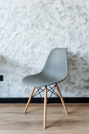A gray chair with elegant wooden legs is positioned comfortably in front of a solid concrete wall, creating a striking contrast between the two materials. The design is modern and stylishの写真素材