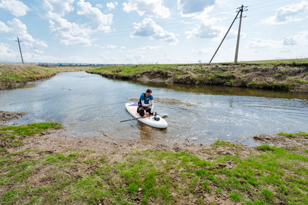 A paddleboarder comfortably sits on a gentle stream, completely surrounded by vibrant, lush greenery, reflecting a perfect serene outdoor experience on a bright sunny dayの写真素材