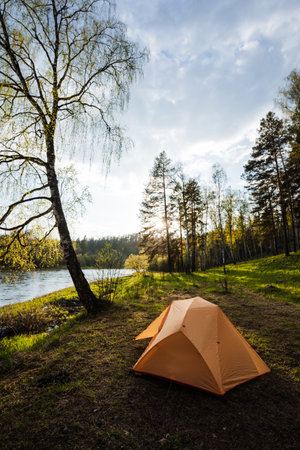 A cozy, vibrant orange tent is beautifully nestled by a tranquil river, completely surrounded by lush greenery and towering trees, making it the perfect spot for camping and outdoor adventuresの写真素材