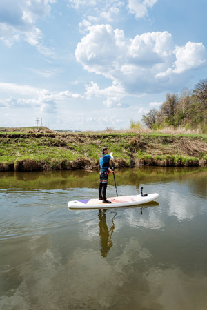 A skilled paddleboarder glides across a tranquil river, flanked by lush greenery, under a bright blue sky and fluffy clouds, creating a perfect scene for relaxation and nature connectionの写真素材