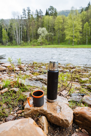 A sleek black thermos and a vibrant orange cup rest on rocky terrain, with a serene river and lush trees in the background, creating an idyllic scene for enjoying coffee or tea in natureの写真素材
