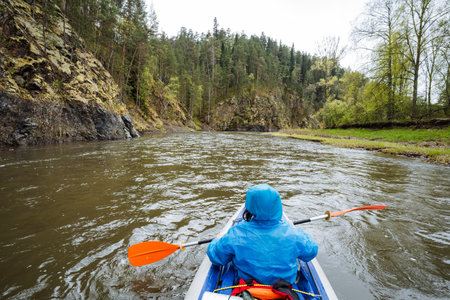 A skilled kayaker in a vibrant blue raincoat expertly navigates serene waters, surrounded by lush...