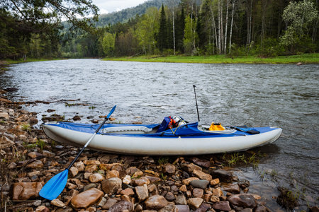 A kayak rests on a rocky river shore, surrounded by vibrant greenery and distant mountains, perfect for adventure enthusiasts and nature lovers seeking exploration and relaxation in the outdoorsの写真素材
