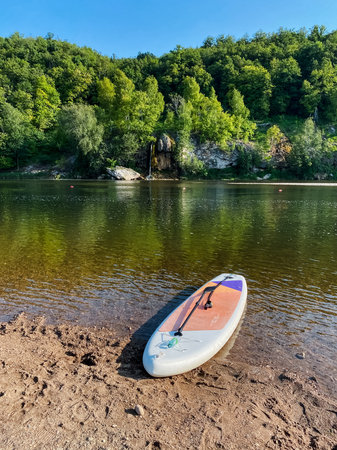 A paddle board is resting peacefully on the tranquil shore of a picturesque lake, surrounded by nature, inviting people to enjoy recreation during the warm, sunny summer days aheadの写真素材