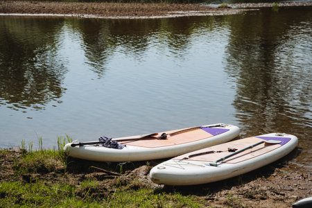 Two vibrant StandUp Paddleboards rest on the sandy shore of a tranquil river, inviting adventurers...