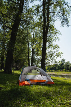 A peaceful camping scene features a colorful tent on a grassy bank, surrounded by tall trees next to a calm riveran ideal spot for outdoor enthusiasts and nature lovers seeking wildernessの写真素材