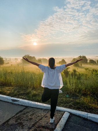 A woman joyfully stands on one leg in a vast green field, arms wide open, embracing the stunning view all around her while the warmth of the sun gently kisses her skin, radiating pure happinessの写真素材