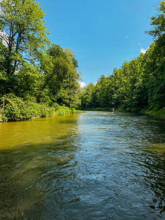 On a beautiful and sunny day, a serene river flows gracefully, bordered by lush trees that provide shade and enhance the stunning natural environment surrounding the peaceful watercourseの写真素材