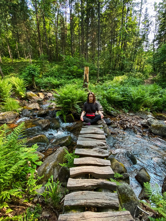 A woman is peacefully sitting on a rustic wooden bridge that spans over a gentle stream flowing through the serene woods surrounding her. This tranquil setting is perfect for relaxation and enjoymentの写真素材