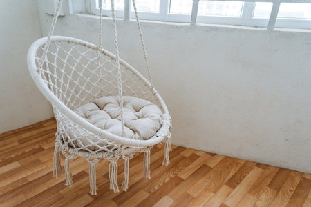 A beautiful white hanging chair is gracefully positioned on a wooden floor, directly in front of a large window that allows natural light to flood the space. It creates a cozy atmosphereの写真素材