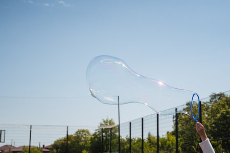 A colorful soap bubble floats against a clear blue sky at a sunny playground, evoking joy and...
