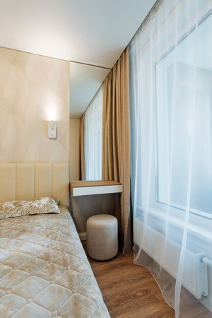 This is a cozy bedroom that features a comfortable bed, a stylish ottoman, a decorative mirror, and a lovely window that lets in natural light, creating a warm and inviting atmosphereの写真素材